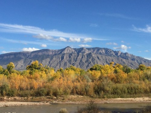 Scenic view of mountains with a blue sky, surrounded by trees with autumn foliage along a riverbank.