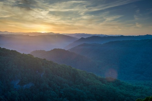 A serene landscape of rolling hills and valleys at sunset, with soft mist and a colorful sky.