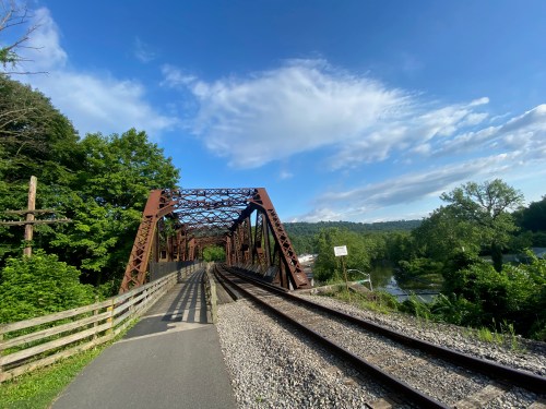 A rusty metal bridge spans a river, with a bike path alongside and lush greenery in the background under a blue sky.