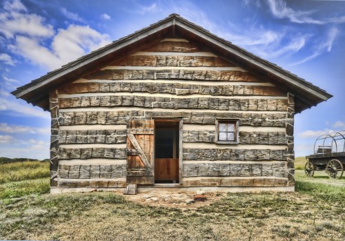A rustic log cabin with a wooden door and window, set against a blue sky and grassy landscape.
