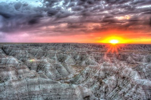 Sunset over rugged Badlands terrain, with dramatic clouds and layered rock formations illuminated by warm light.