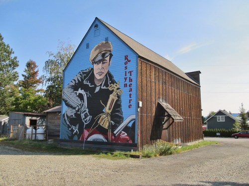 A large mural on a barn depicts a person in a cap holding a trophy, with "Ros Theatre" written above.