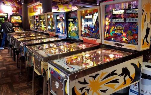 A row of colorful pinball machines in a vibrant arcade setting, with players enjoying the games.