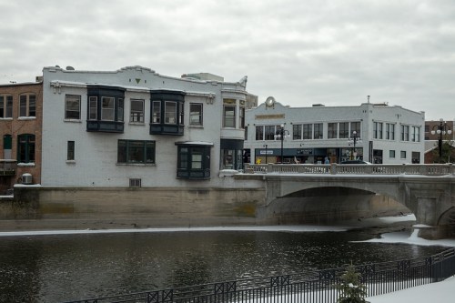A snowy riverside view featuring historic buildings and a bridge over a calm waterway.