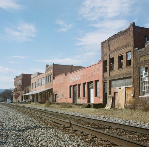 Abandoned brick buildings line a railway track under a clear blue sky, showcasing a forgotten industrial area.