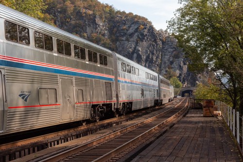 A silver train with red and blue stripes travels along tracks near a rocky landscape and a dark tunnel entrance.