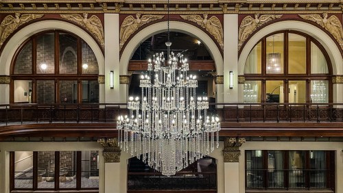 A grand chandelier hangs from a beautifully ornate ceiling, surrounded by arched windows and elegant architectural details.