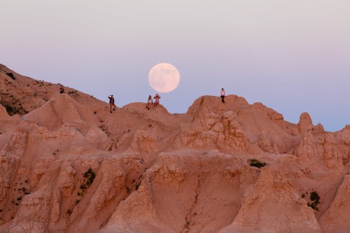 A full moon rises over rugged, sandy hills with people exploring the landscape at dusk.