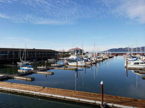 A serene marina with boats docked, reflecting blue skies and mountains in calm water.