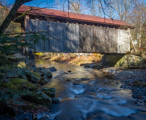 A wooden covered bridge spans a flowing creek, surrounded by trees and autumn foliage under a clear blue sky.