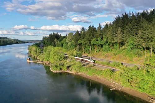 A scenic river view with lush green trees, a train along the bank, and a partly cloudy sky.
