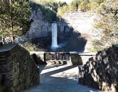 A stone staircase leads to a viewpoint of a waterfall surrounded by lush greenery and rocky cliffs.