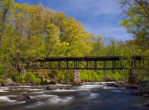 A rustic wooden bridge spans a flowing river, surrounded by lush green trees under a clear blue sky.