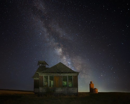 An abandoned house under a starry sky, with the Milky Way visible and a silhouette of a grain silo nearby.