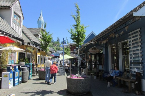 A sunny street scene with shops, outdoor seating, and people enjoying the vibrant atmosphere. Green trees and umbrellas add color.