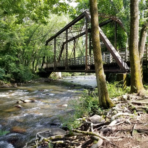 A rustic metal bridge spans a flowing river, surrounded by lush green trees and rocky riverbanks.