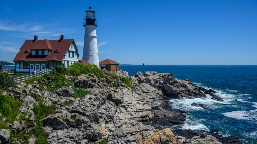 A scenic lighthouse beside a rocky shore, with a charming house and clear blue sky in the background.