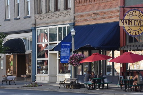 A quaint street scene featuring shops with awnings, outdoor seating, and red umbrellas in a small town.