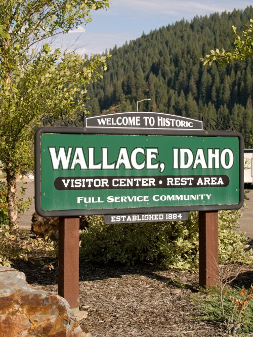 Welcome sign for Wallace, Idaho, featuring "Visitor Center" and "Rest Area," surrounded by greenery. Established 1884.