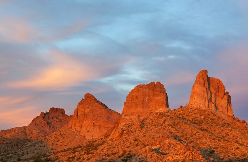Sunset over rugged mountains, casting warm orange hues against a colorful sky with soft clouds.