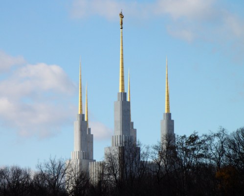 A tall, modern building with four golden spires against a blue sky, surrounded by trees.