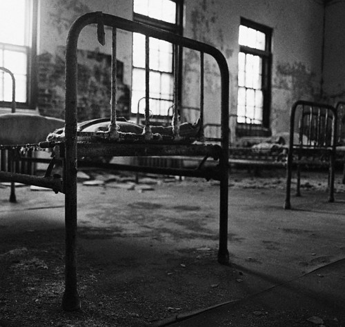 Abandoned room with rusty metal beds, dust-covered floors, and peeling walls, evoking a sense of decay and neglect.
