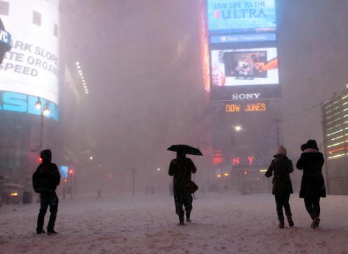 People walking in a snowy Times Square, New York, with large illuminated billboards in the background.