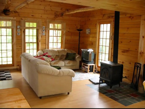 Cozy wooden living room with a sofa, TV, and a wood stove, featuring large windows and natural light.