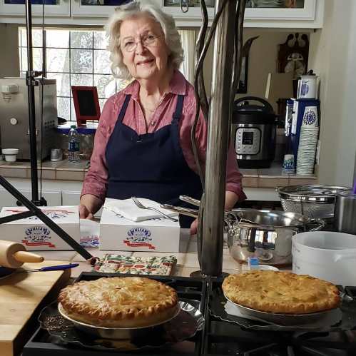 A smiling woman in an apron stands in a kitchen with two freshly baked pies on the counter.
