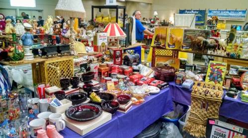 A bustling flea market table filled with vintage items, dishes, toys, and collectibles, with shoppers in the background.