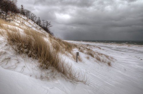 A snowy beach scene with tall grass, dunes, and dark, cloudy skies over a turbulent lake.