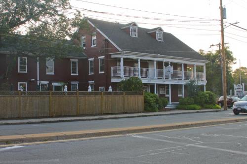 A red two-story building with a porch, surrounded by greenery and a wooden fence, along a street at sunset.