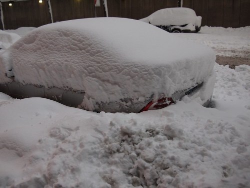A car completely covered in thick snow, with only the outline visible, surrounded by a snowy landscape.