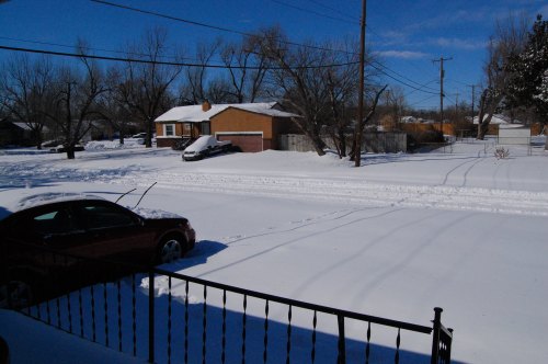A snowy residential street with a parked car and houses in the background under a clear blue sky.
