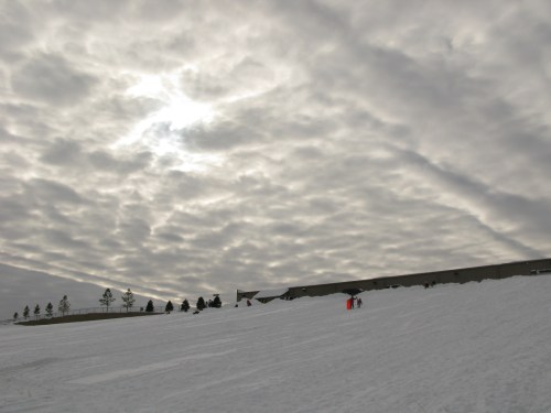 A snowy landscape under a cloudy sky, with a few people in the distance near a building.