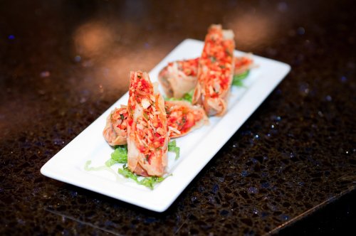 A white plate with three spring rolls filled with colorful vegetables, garnished with lettuce on a dark surface.