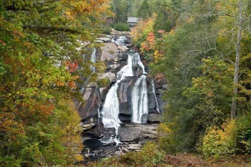 A scenic waterfall surrounded by vibrant autumn foliage and rocky terrain, with a cabin visible in the background.