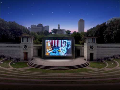 An outdoor amphitheater at dusk, featuring a large screen displaying a film, surrounded by city skyline and trees.