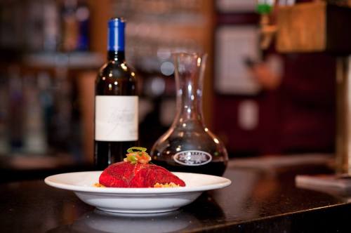 A plate of red pasta topped with herbs, accompanied by a bottle of wine and a decanter on a bar counter.