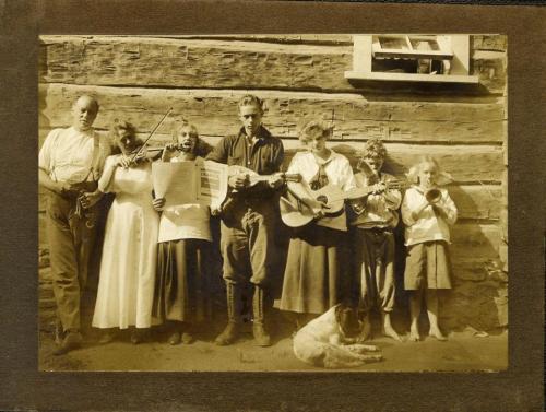 A sepia-toned photo of a family band with instruments, standing in front of a log cabin, with a dog sitting nearby.