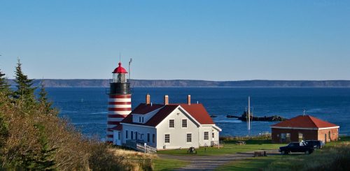 A striped lighthouse beside a white house, overlooking the ocean and distant cliffs under a clear blue sky.