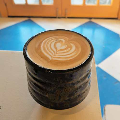 A latte in a black ceramic cup, featuring a heart-shaped foam design, sits on a blue and white patterned table.