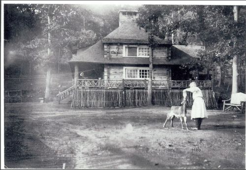 A historic black-and-white photo of a woman in a white dress with a dog in front of a log cabin surrounded by trees.
