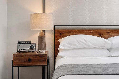 A cozy bedroom scene featuring a wooden bed, bedside table with a lamp, clock radio, and glasses.