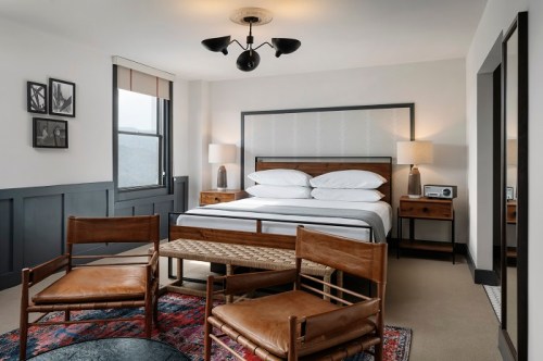 A modern hotel room featuring a queen bed, two wooden chairs, a table, and stylish decor with natural light.