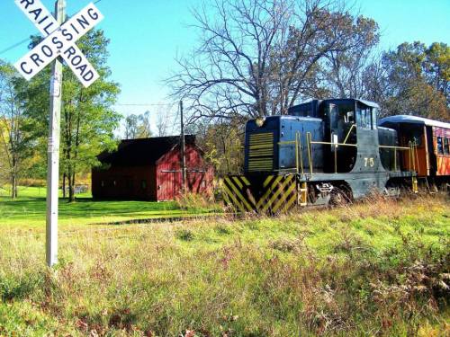A black train engine near a railroad crossing sign, with a red barn and trees in the background.
