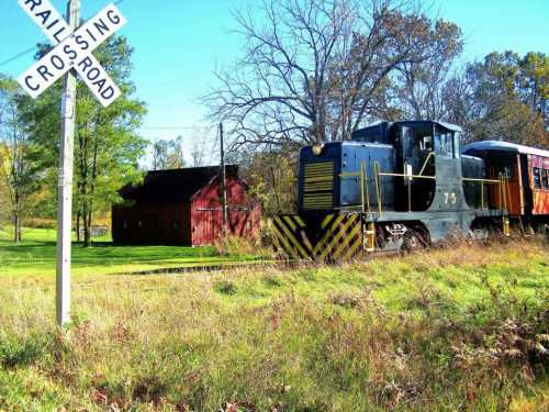 A black train engine near a railroad crossing sign, with a red barn and trees in the background.