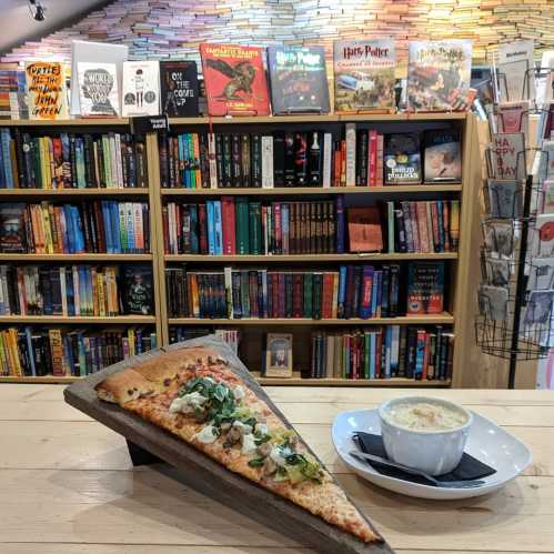 A slice of pizza and a bowl of soup on a wooden table, with a colorful bookshelf filled with books in the background.