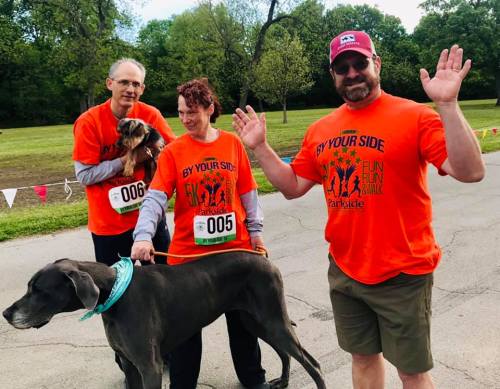 Two people in orange shirts with dogs, smiling and waving at a park during a fun run event.