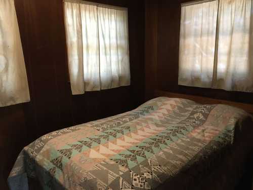 A cozy bedroom with a quilted bedspread and two windows covered with sheer curtains.